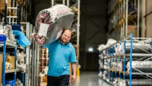 Man in a blue polo shirt carrying a large, wrapped package on his shoulder in a warehouse aisle with shelves and various items.