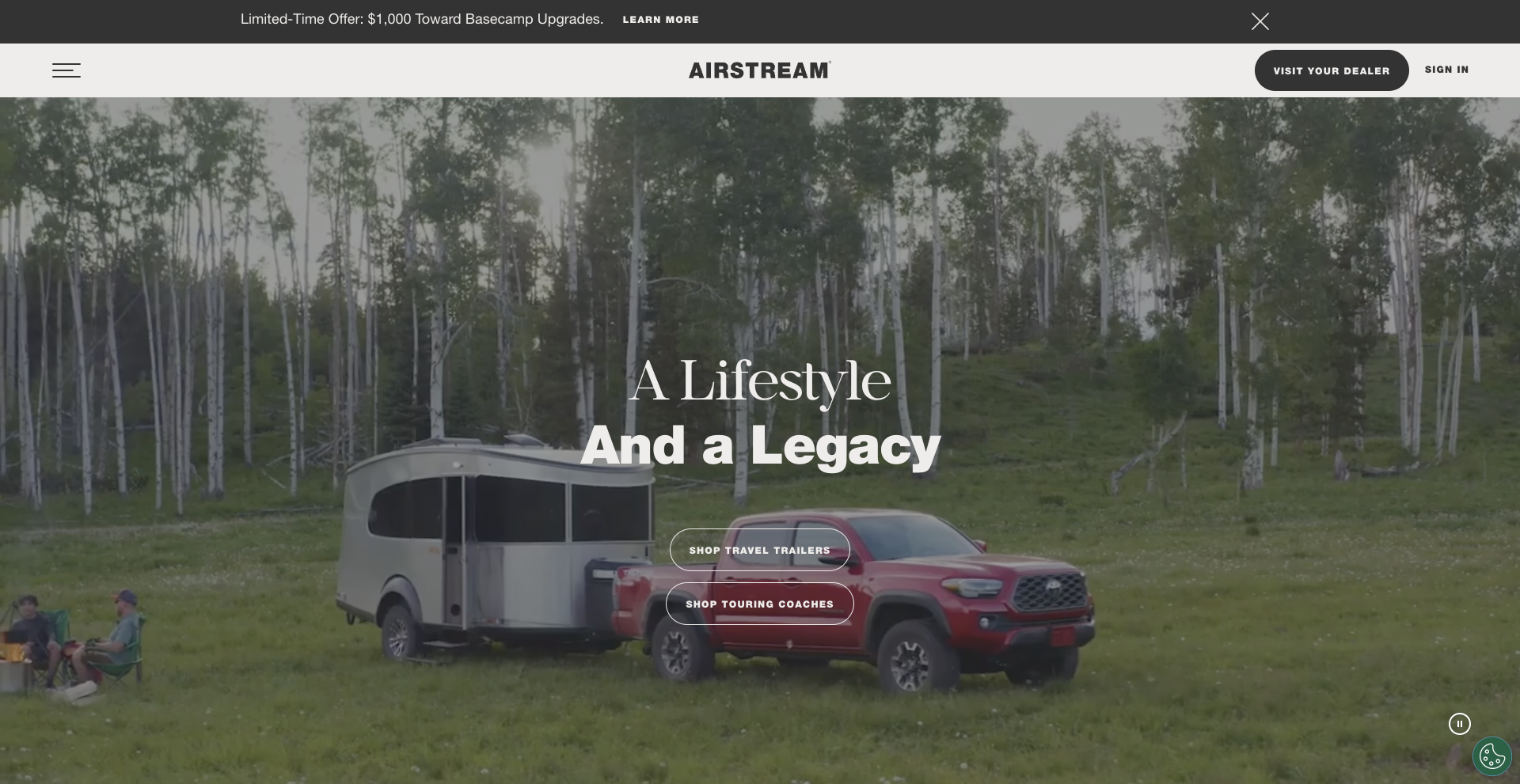 Alt text: "Airstream website homepage banner reading 'A Lifestyle And a Legacy,' showing a silver Airstream Basecamp trailer hitched to a red pickup truck parked in a grassy forest clearing, with a family relaxing at a campsite in the background. A promotional bar at top advertises a limited-time $1,500 Basecamp upgrade offer, alongside navigation and 'Visit Your Dealer' and 'Sign In' buttons.