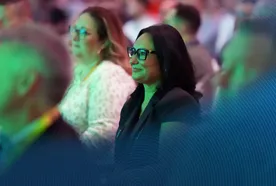 Audience members, including a woman in glasses, attentively watching at an event. The scene has a green lighting effect.