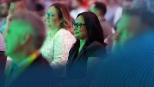 Audience members, including a woman in glasses, attentively watching at an event. The scene has a green lighting effect.