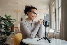 Woman with glasses smiling at smartphone on tripod, sitting at a table in a bright room with plants and large windows.