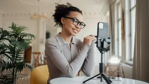 Woman with glasses smiling at smartphone on tripod, sitting at a table in a bright room with plants and large windows.
