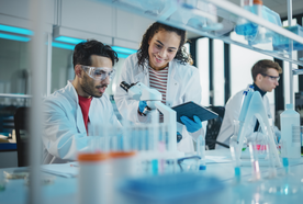 Scientists in a lab, one using a microscope, the other holding a tablet, both wearing lab coats and goggles; blurred lab equipment in foreground.