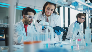 Scientists in a lab, one using a microscope, the other holding a tablet, both wearing lab coats and goggles; blurred lab equipment in foreground.