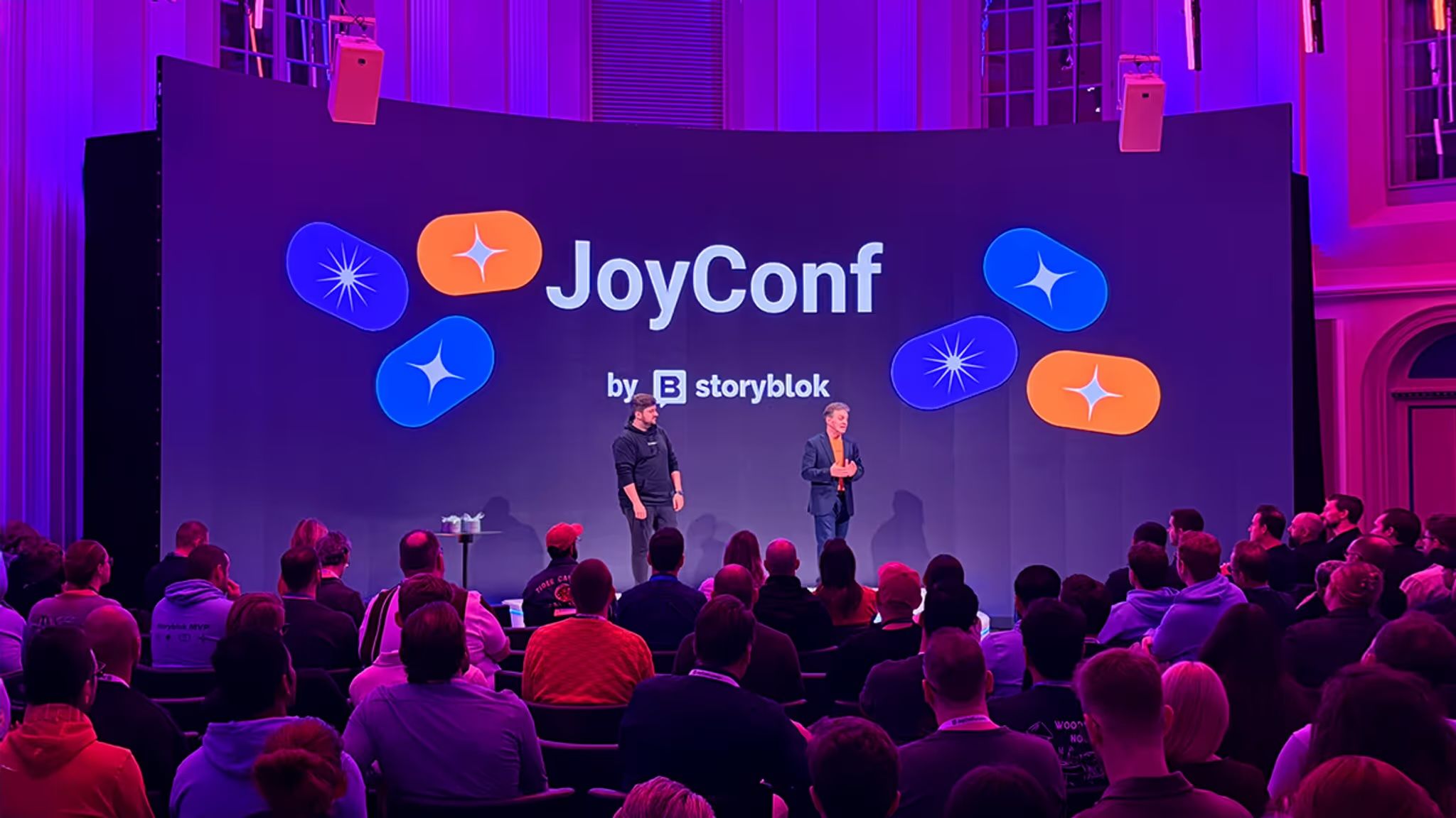 Two people on stage at JoyConf by Storyblok, with a seated audience. The backdrop is purple with logo designs.
