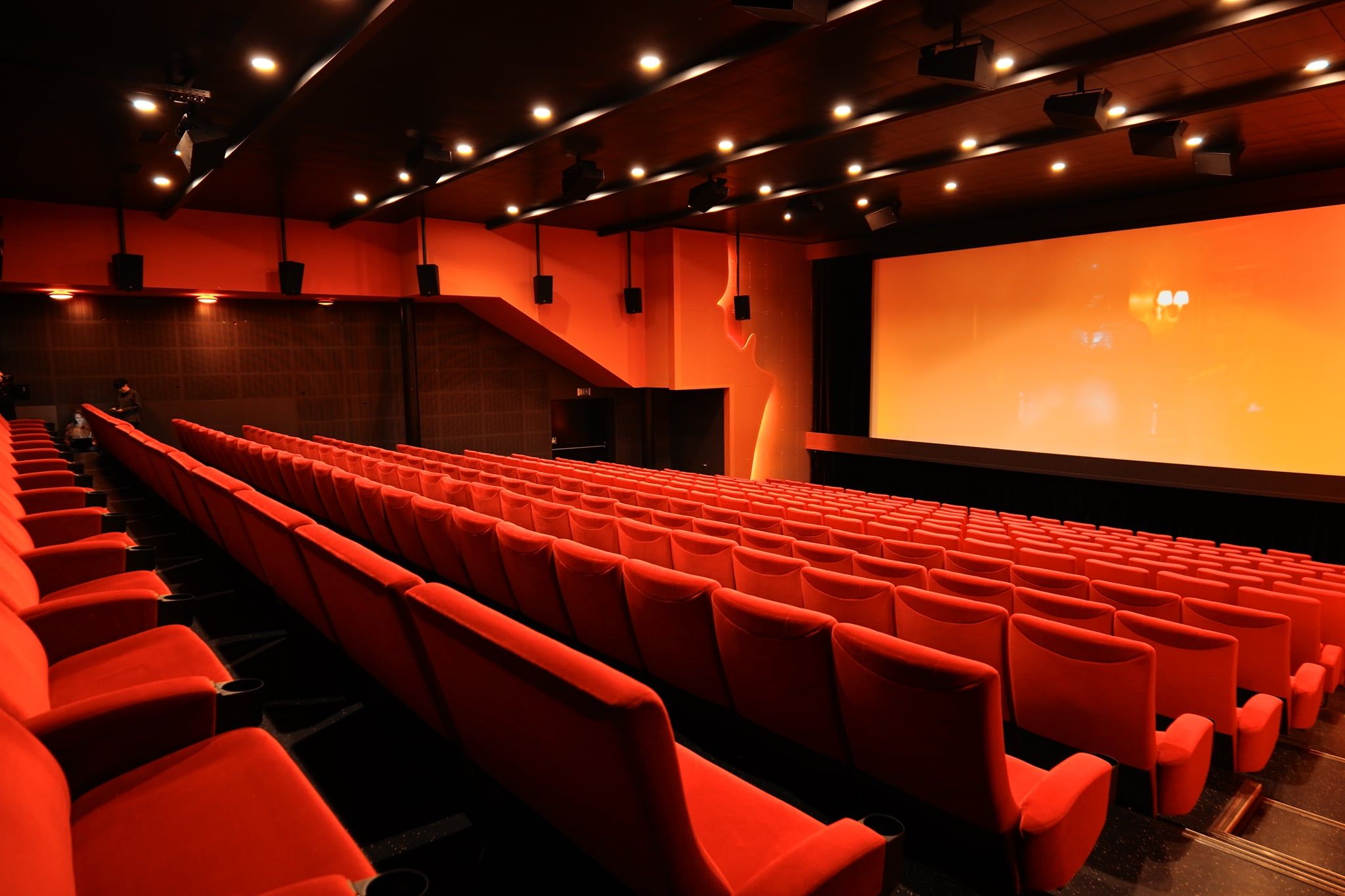 Empty cinema auditorium with rows of red seats facing a large screen. Dim lighting and speakers are visible on the walls and ceiling.