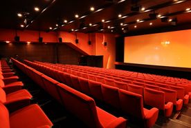 Empty cinema auditorium with rows of red seats facing a large screen. Dim lighting and speakers are visible on the walls and ceiling.