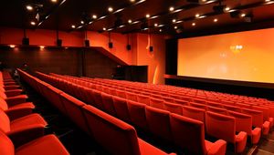 Empty cinema auditorium with rows of red seats facing a large screen. Dim lighting and speakers are visible on the walls and ceiling.