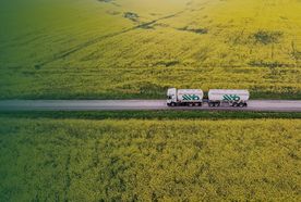 Aerial view of a tanker truck driving down a road through vast yellow fields, possibly canola, under a green-tinted sky.