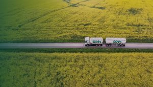 Aerial view of a tanker truck driving down a road through vast yellow fields, possibly canola, under a green-tinted sky.