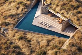 Aerial view of a modern house integrated into a hillside with a triangular pool, stone walls, and outdoor seating, surrounded by dry grassland.