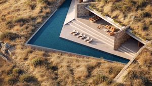 Aerial view of a modern house integrated into a hillside with a triangular pool, stone walls, and outdoor seating, surrounded by dry grassland.