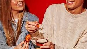 A smiling couple enjoys spicy chicken wings together at a small table, set against a bright red background.