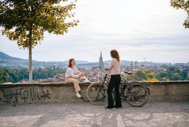 Two people converse joyfully on a scenic overlook, with a bike nearby and a picturesque cityscape in the background.