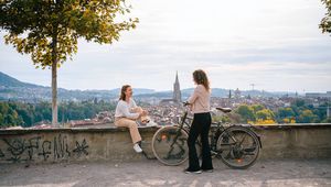 Two people converse joyfully on a scenic overlook, with a bike nearby and a picturesque cityscape in the background.