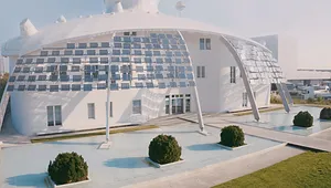 Futuristic white building with solar panels and curved design, surrounded by a shallow pool and neatly trimmed bushes.