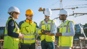 Four construction workers in safety gear discuss plans on a tablet at a building site with cranes in the background.