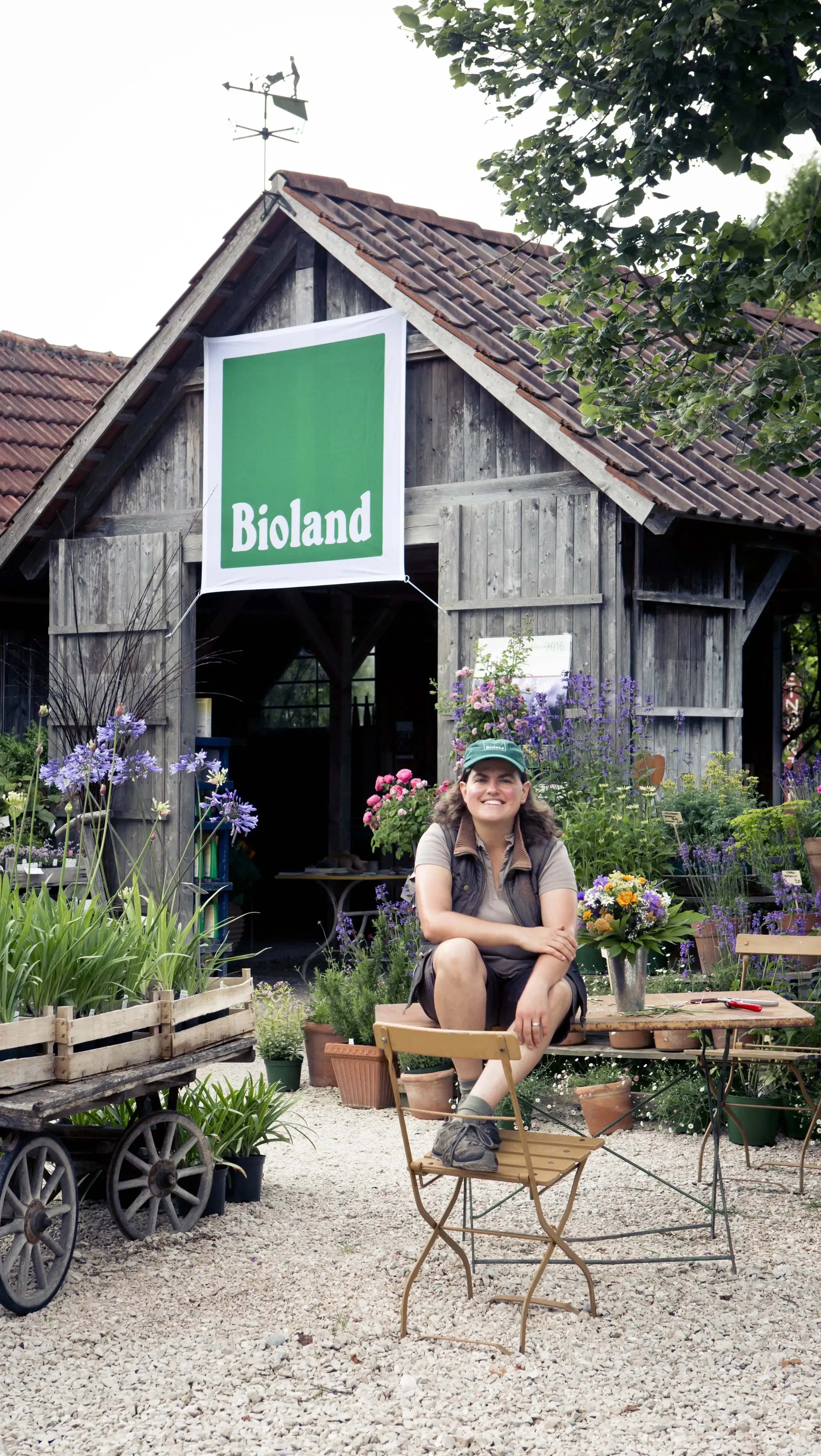 A person sits smiling in front of a rustic wooden farm building with a large green “Bioland” sign hanging above the entrance. The scene is surrounded by flowers, potted plants, and wooden carts filled with greenery, creating a cozy organic farm atmosphere.