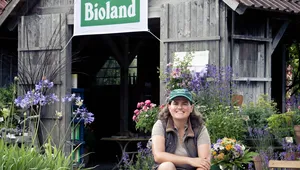 A person sits smiling in front of a rustic wooden farm building with a large green “Bioland” sign hanging above the entrance. The scene is surrounded by flowers, potted plants, and wooden carts filled with greenery, creating a cozy organic farm atmosphere.