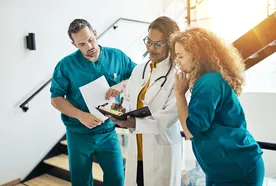 Three medical professionals in scrubs and a lab coat discuss notes in a bright, modern hallway.