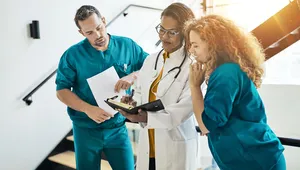 Three medical professionals in scrubs and a lab coat discuss notes in a bright, modern hallway.