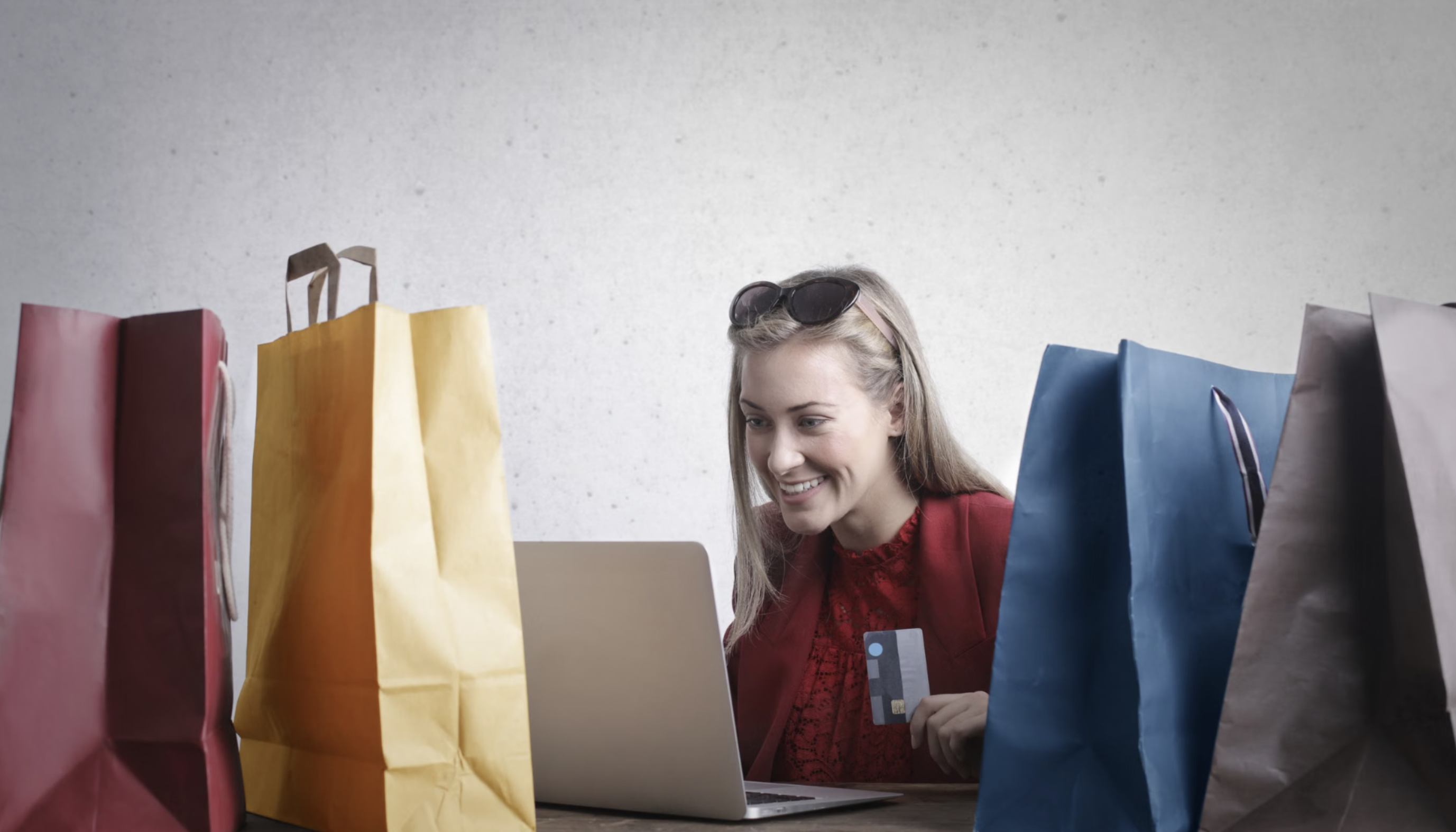 Smiling woman shopping online with a credit card, surrounded by colorful shopping bags, sitting at a table with a laptop.