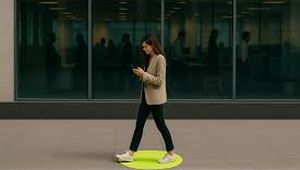 Woman walking on a sidewalk, looking at her phone, with a green circle on the ground, in front of a glass building.
