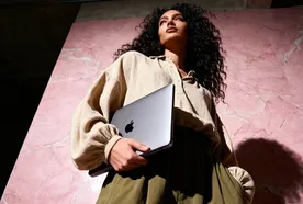 A person with curly hair holds a laptop against a pink marble wall, dressed in a beige shirt and olive green skirt, looking upwards.