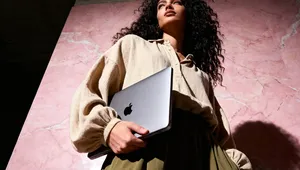 A person with curly hair holds a laptop against a pink marble wall, dressed in a beige shirt and olive green skirt, looking upwards.