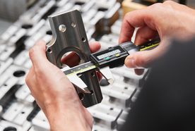 Hands measuring a metal part with a digital caliper, with other machined parts in the background.