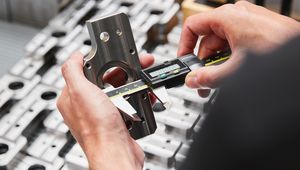 Hands measuring a metal part with a digital caliper, with other machined parts in the background.