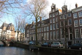 Canal in Amsterdam lined with historic brick buildings, arched bridge, and parked cars; bare trees and a church in the background.