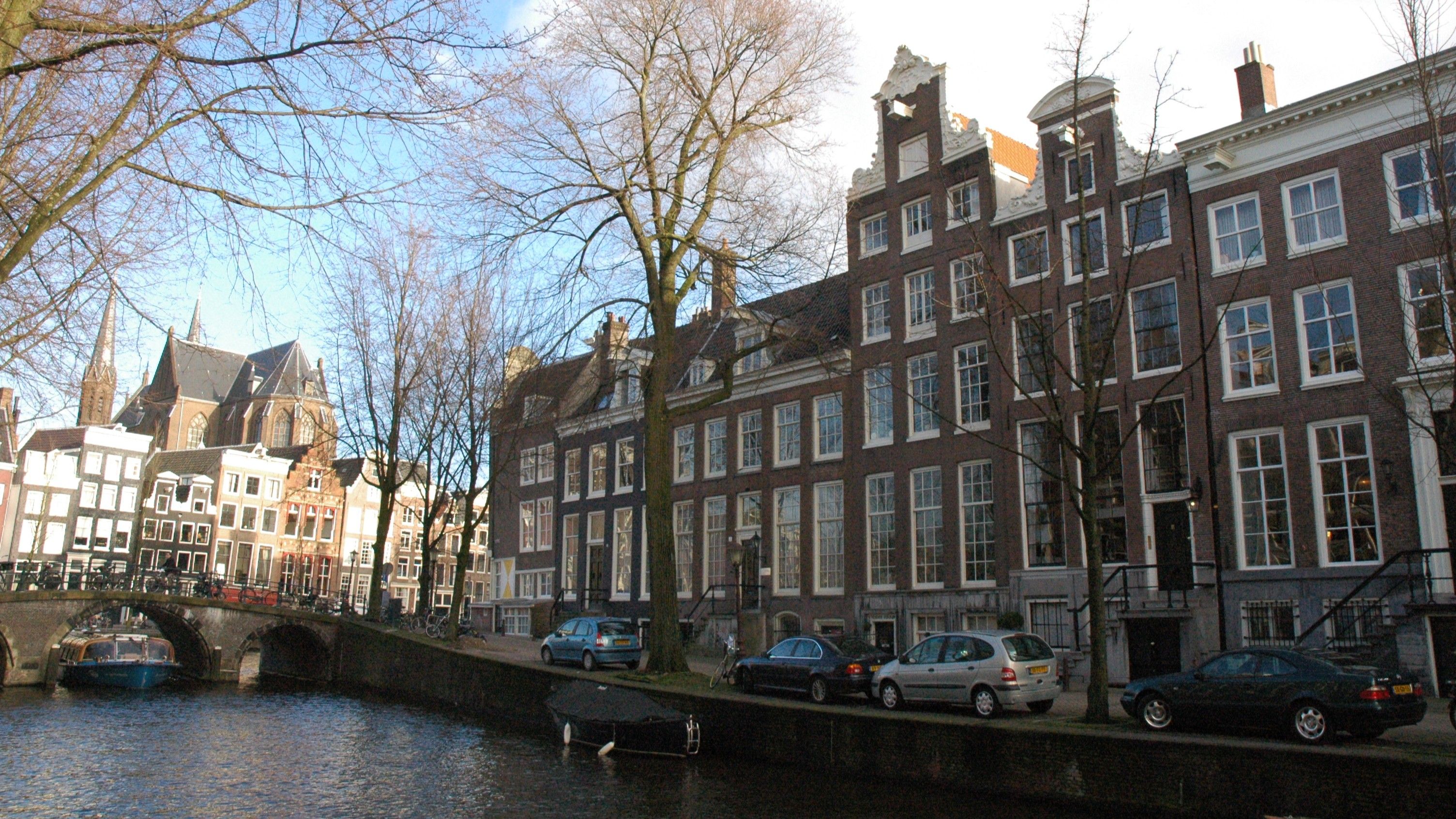Canal in Amsterdam lined with historic brick buildings, arched bridge, and parked cars; bare trees and a church in the background.