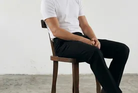 Man in a white t-shirt and black pants sitting on a wooden chair against a plain background, looking to the side.