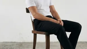 Man in a white t-shirt and black pants sitting on a wooden chair against a plain background, looking to the side.