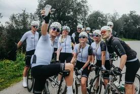 A group of cyclists in matching gear take a selfie on a rural road, smiling and posing with their bikes.