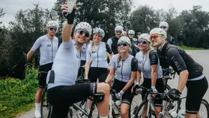A group of cyclists in matching gear take a selfie on a rural road, smiling and posing with their bikes.