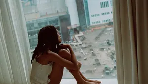 A person with braided hair sits on a windowsill, looking out at a cityscape with buildings and a busy street below.