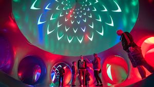 People standing inside an illuminated inflatable dome tunnel, colorful red, green and blue lights with spiral, petal-like projections on the ceiling.
