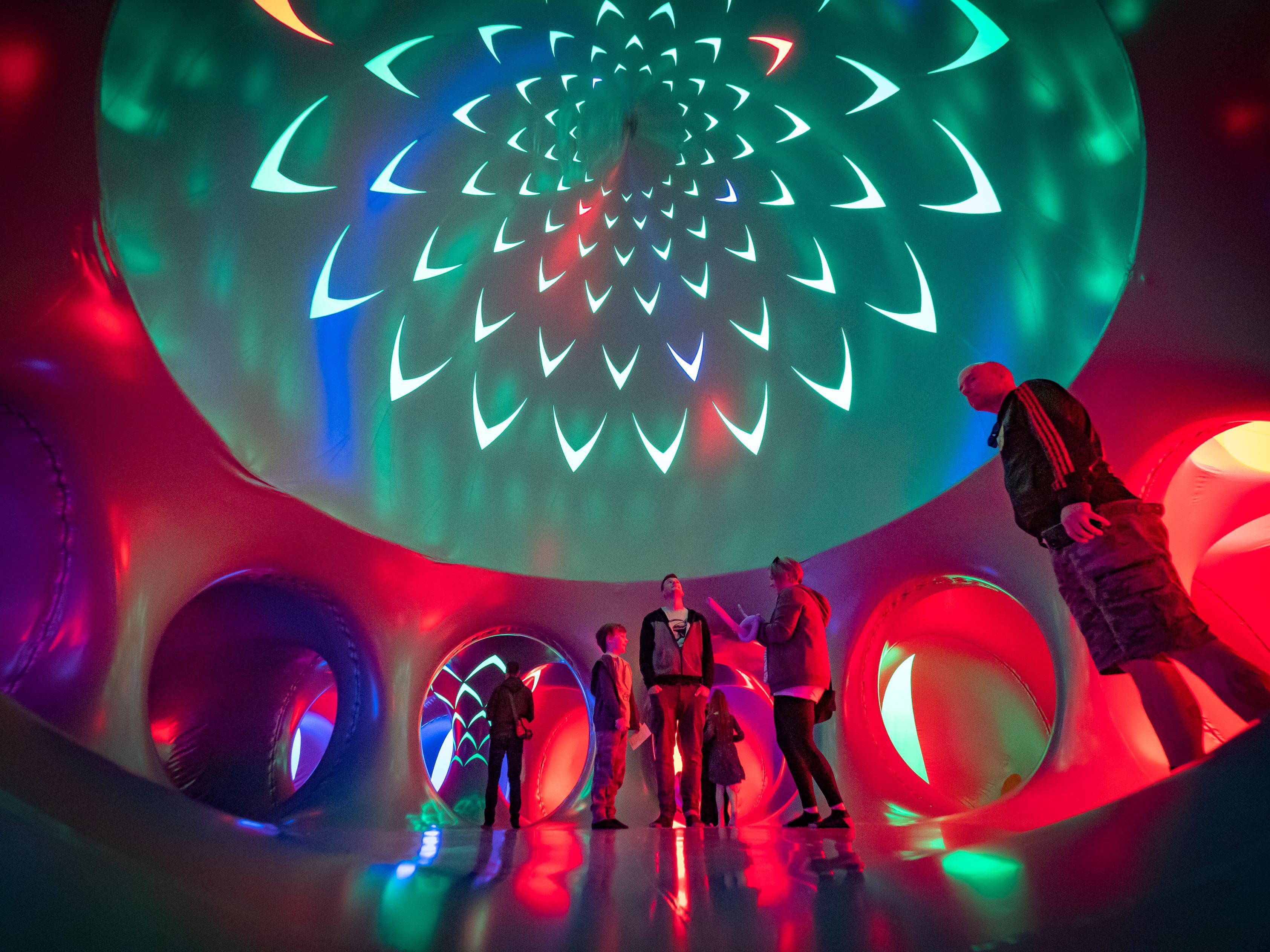 People standing inside an illuminated inflatable dome tunnel, colorful red, green and blue lights with spiral, petal-like projections on the ceiling.