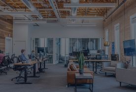 Modern open-plan office with people working at desks, exposed brick walls, large windows, and a central seating area with a plant on a table.