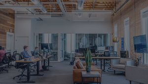 Modern open-plan office with people working at desks, exposed brick walls, large windows, and a central seating area with a plant on a table.