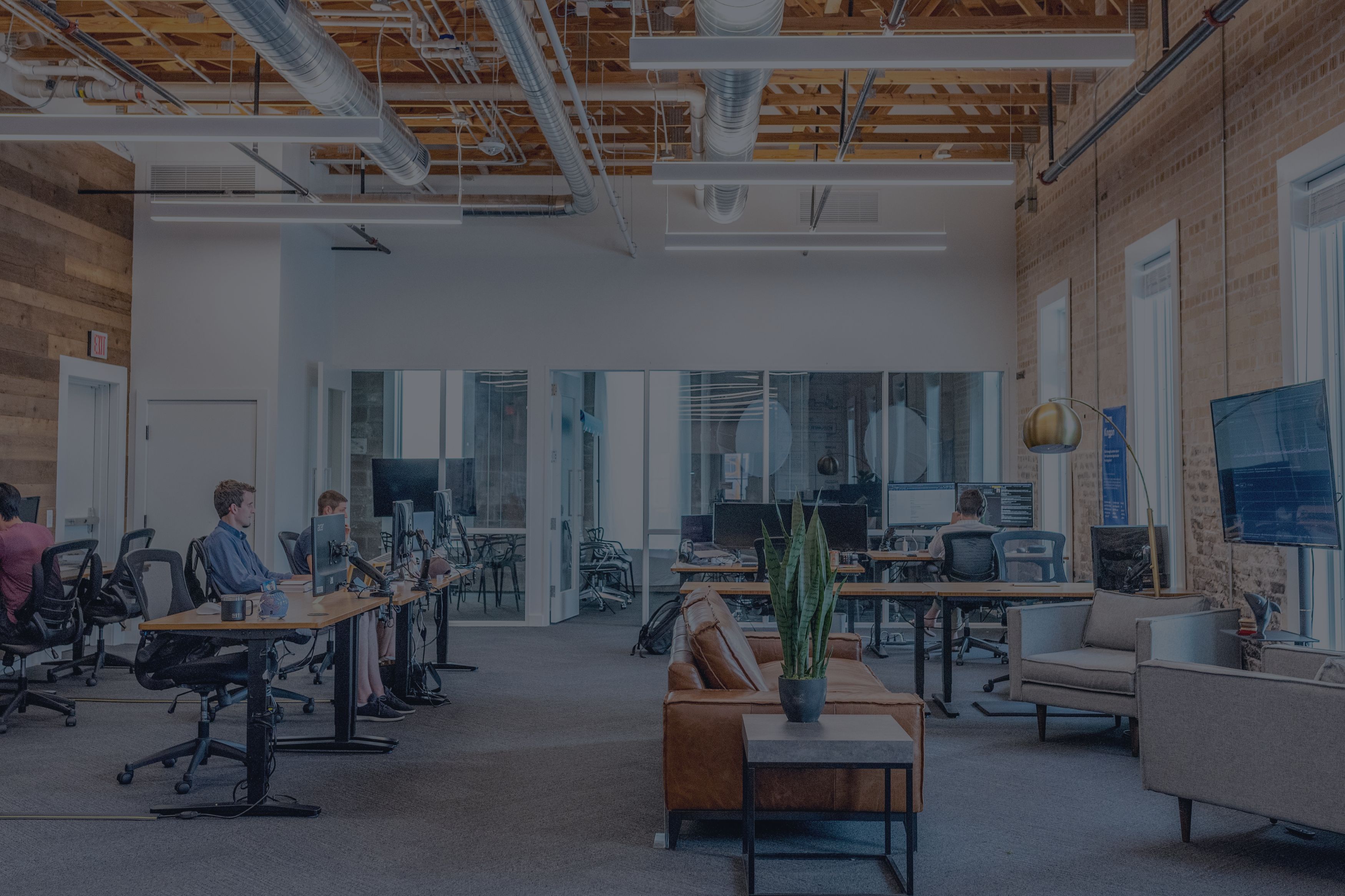Modern open-plan office with people working at desks, exposed brick walls, large windows, and a central seating area with a plant on a table.