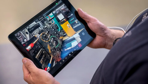 A person holds a tablet displaying a close-up image of industrial machinery. The focus is on detailed engineering components. The tone is technological and focused.