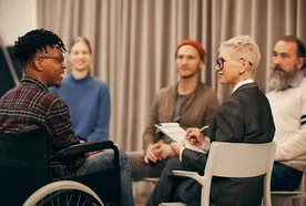 A group of five people in a discussion circle. One person in a wheelchair smiles while a facilitator with a notepad takes notes.