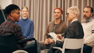A group of five people in a discussion circle. One person in a wheelchair smiles while a facilitator with a notepad takes notes.