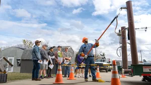 Worker in protective gear demonstrates electrical safety to a group, using a pole to handle equipment, with safety cones marking the area.