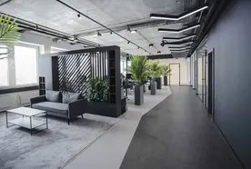 Modern office with gray sofa, black coffee table, and green plants. Open space design with glass partitions and geometric ceiling lights.