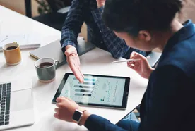 Two professionals discuss data on a tablet at a desk, surrounded by coffee cups and a laptop.