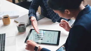 Two professionals discuss data on a tablet at a desk, surrounded by coffee cups and a laptop.